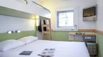 Habitación de hotel con cama, escritorio con ordenador, maleta y ventana con vistas a edificios.