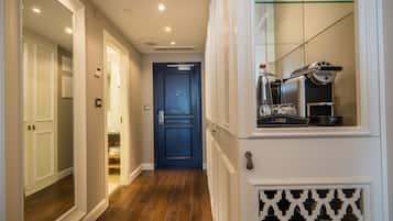 Un pasillo con suelo de madera, paredes blancas y una puerta azul al final. Se ve un radiador y una máquina de café en la pared derecha.