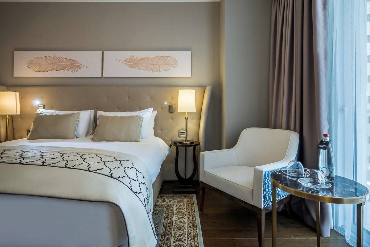 A hotel room with a large bed, two bedside lamps, a chair, a small table with glasses, and a window with curtains.