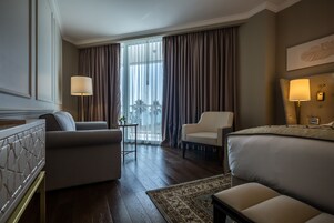Habitación de hotel con una cama grande, dos sillones, una mesita de noche con lámpara y vistas al exterior a través de cortinas translúcidas.