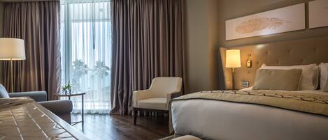 Habitación de hotel con una cama grande, una silla, un escritorio y vistas al exterior a través de amplias ventanas.