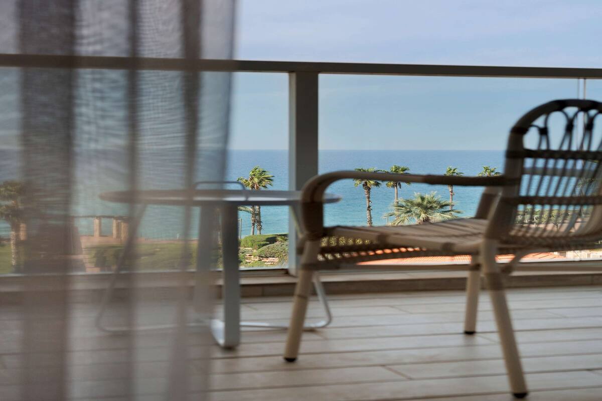 A balcony with a wicker chair and a small table, overlooking a coastal view with palm trees and the sea.