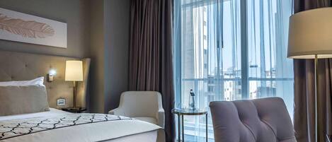 Habitación de hotel con cama, dos sillas, una mesita, lámpara y ventana grande con cortinas.