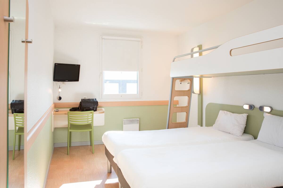 A hotel room with a bunk bed, a desk with a chair, a television mounted on the wall, and a window with blinds.
