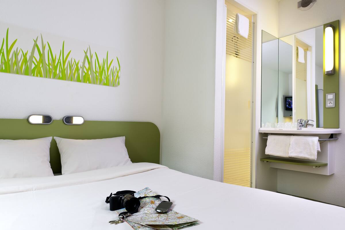 A hotel room with a green headboard, white bedding, a bed with a camera and map, and a bathroom with a mirror and towel rack.