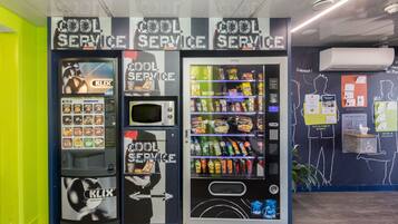 Zona de máquinas expendedoras con un letrero que dice 'Cool Service', una máquina de café K-LIX y un dispensador de bebidas.