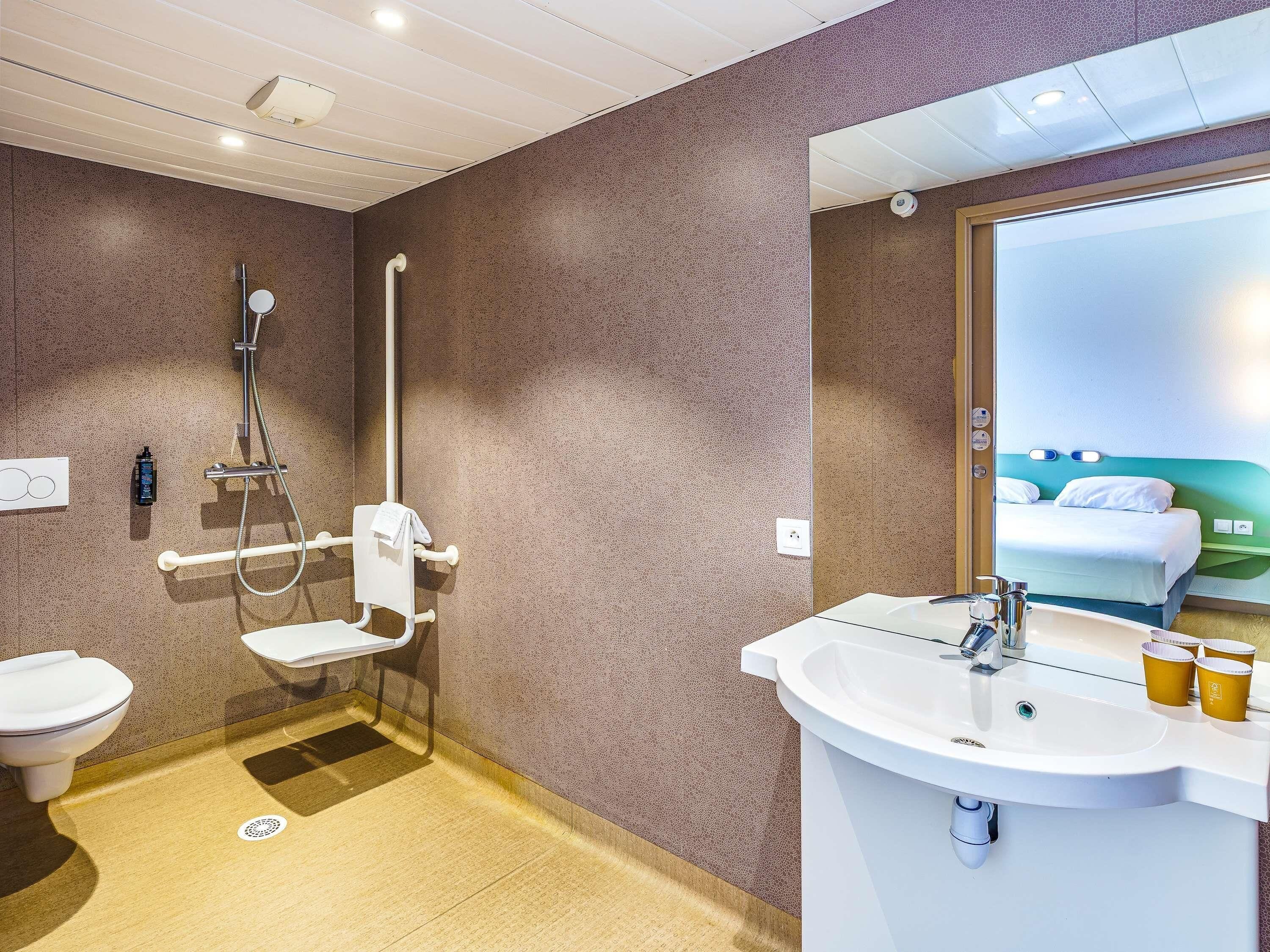 Ein modernes Badezimmer mit Dusche, Toilette und Waschbecken, ausgestattet mit einem großen Spiegel und Blick auf ein Bett im angrenzenden Zimmer.