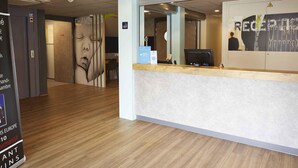 A reception area with a wooden floor, a reception desk, and a wall with a large mirror reflecting the interior. - ibis budget Chalon Sur Saone Nord (Chalon-sur-Saone)