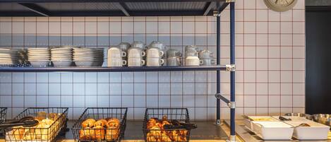 A kitchen area with shelves holding plates, mugs, and baskets of pastries.