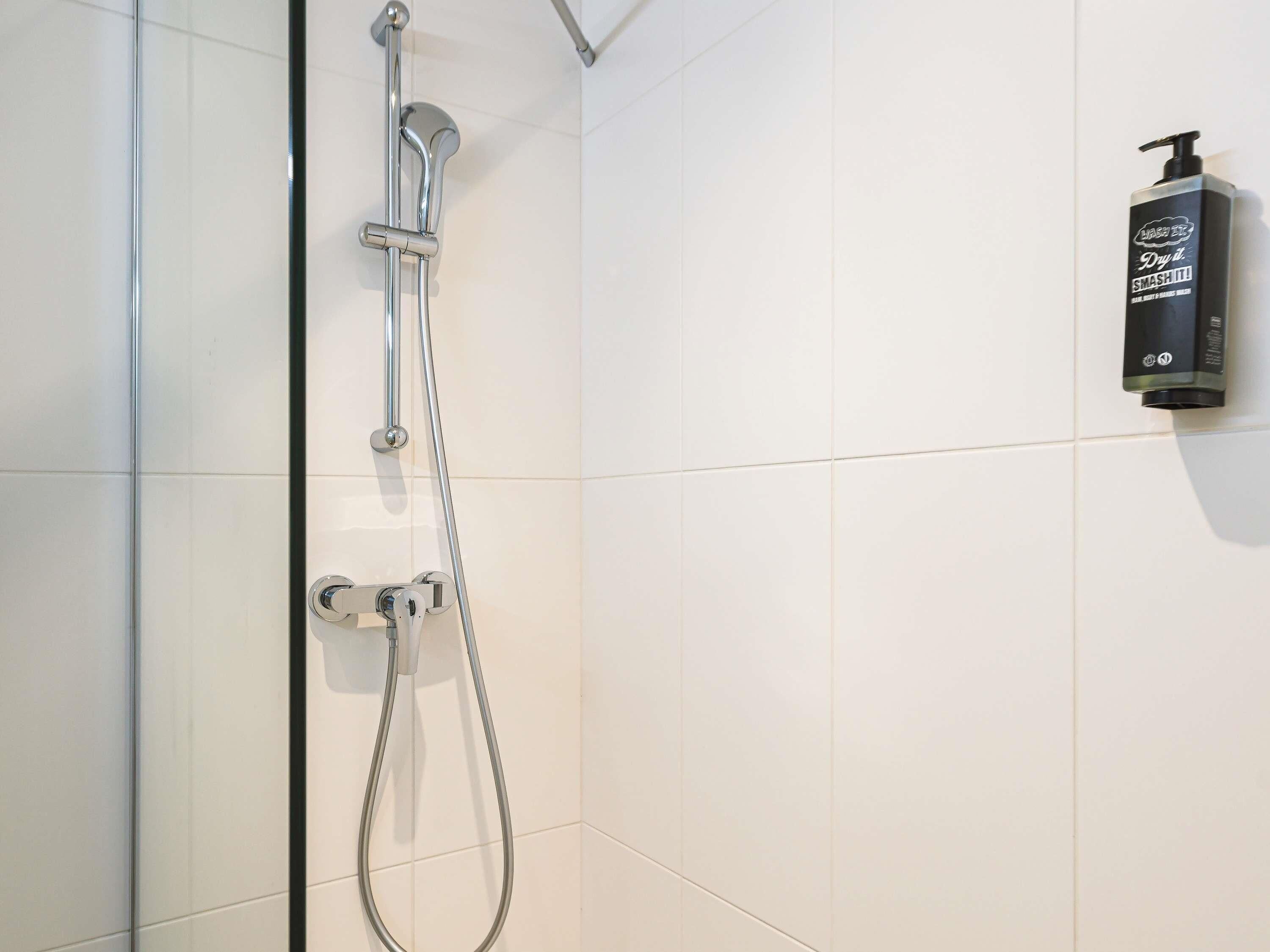 Une douche avec des accessoires chromés et un pommeau de douche à main, un distributeur de savon mural et des murs recouverts de carreaux blancs.