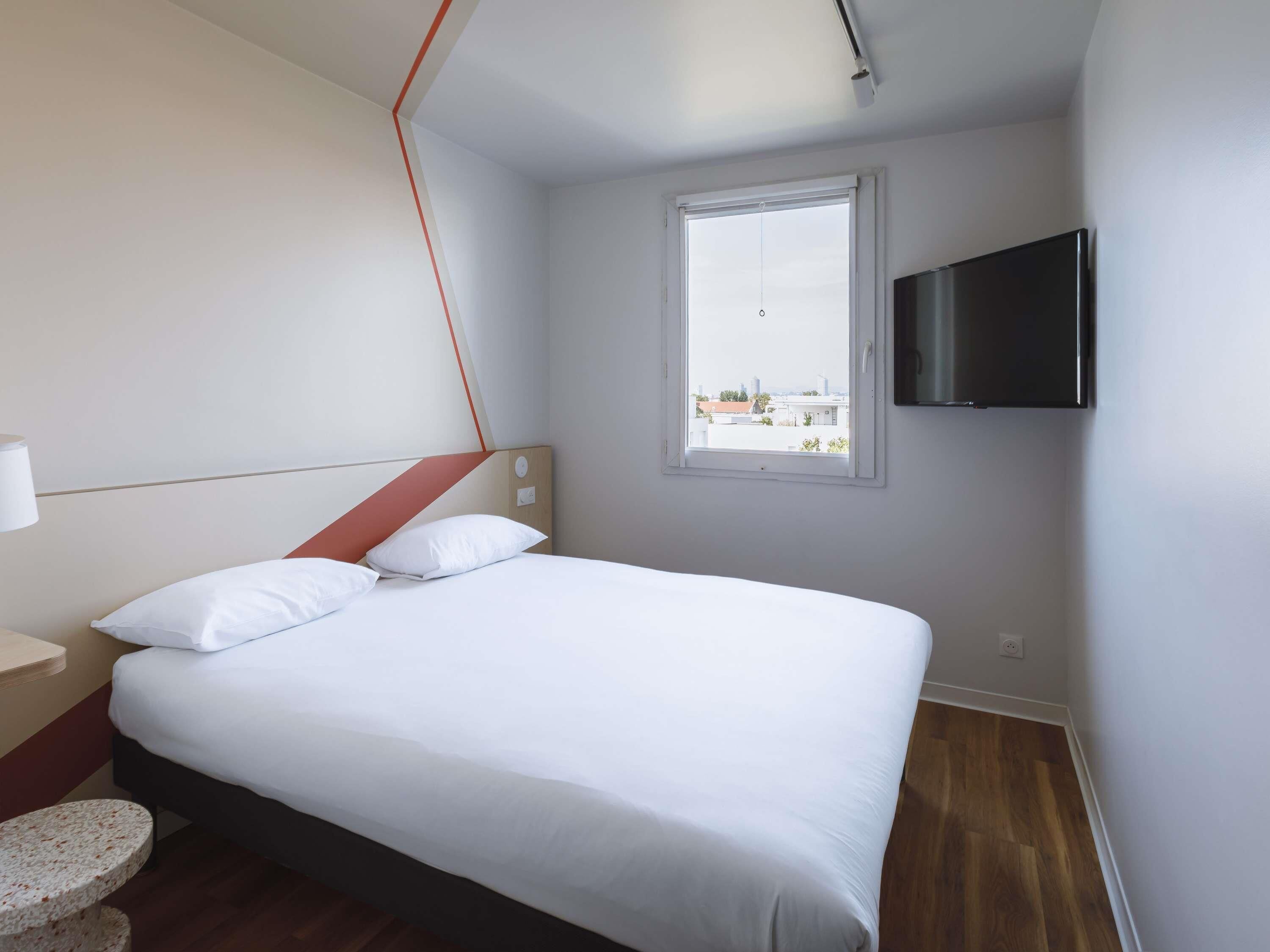 A modern hotel room with a bed, a TV mounted on the wall, and a window with a city view.