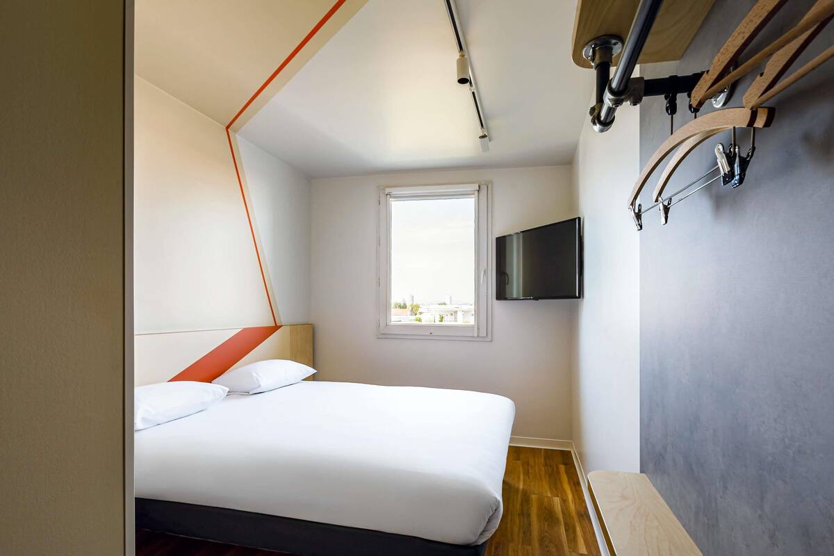 Un dormitorio moderno con una cama, un televisor montado en la pared, una ventana y un banco de madera.