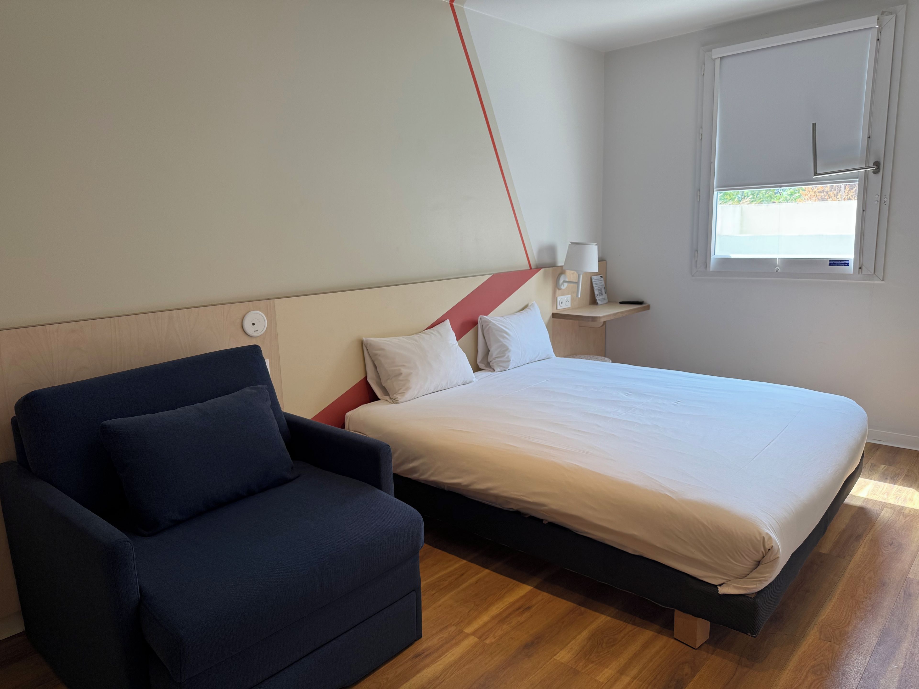 Un dormitorio con cama, mesita de noche, lámpara, ventana y una silla.