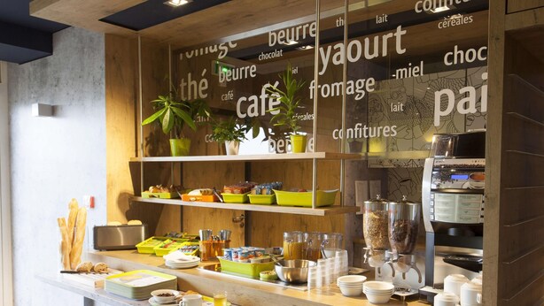 A cafe counter with a coffee machine, various food items, and a plant. - ibis budget Cherbourg La Glacerie (Cherbourg-en-Cotentin)