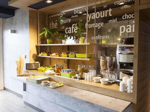 A cafe counter with a coffee machine, various food items, and a plant. - ibis budget Cherbourg La Glacerie (Cherbourg-en-Cotentin)