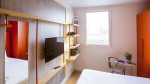 A modern room with a bed, a TV mounted on a wooden shelf, a small table with a plant, and a window with a view. - ibis budget Cherbourg La Glacerie (Cherbourg-en-Cotentin)