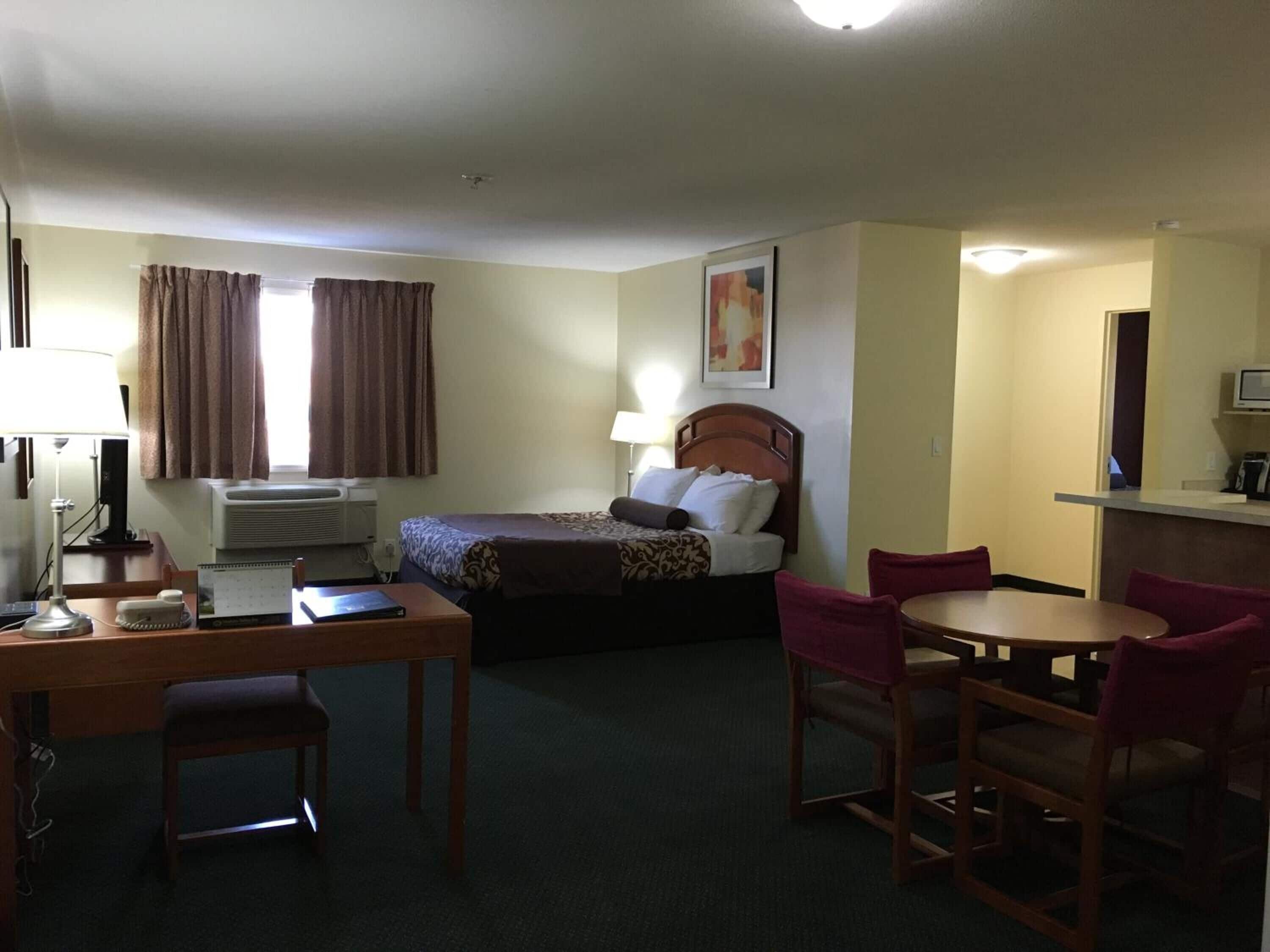 A hotel room with a bed, a desk with a chair, a dining table with chairs, and a television.