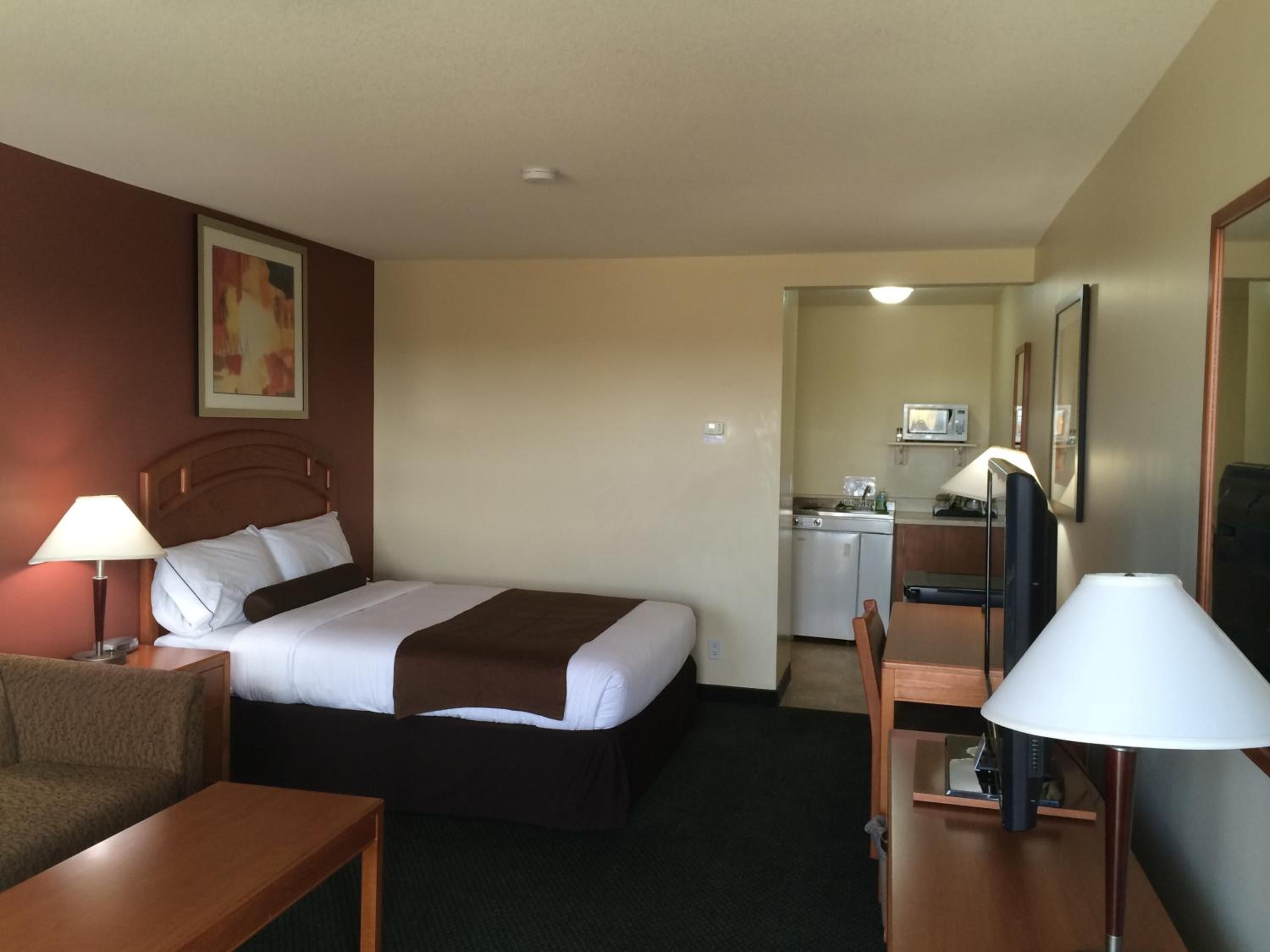 A hotel room with a bed, a desk, a chair, a kitchenette, and a mirror.