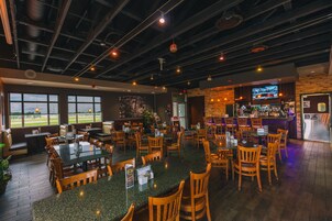 A restaurant with multiple seating areas, including booths and tables. There is a bar area with a TV screen and a counter. The restaurant features large windows and a view of the outdoors.