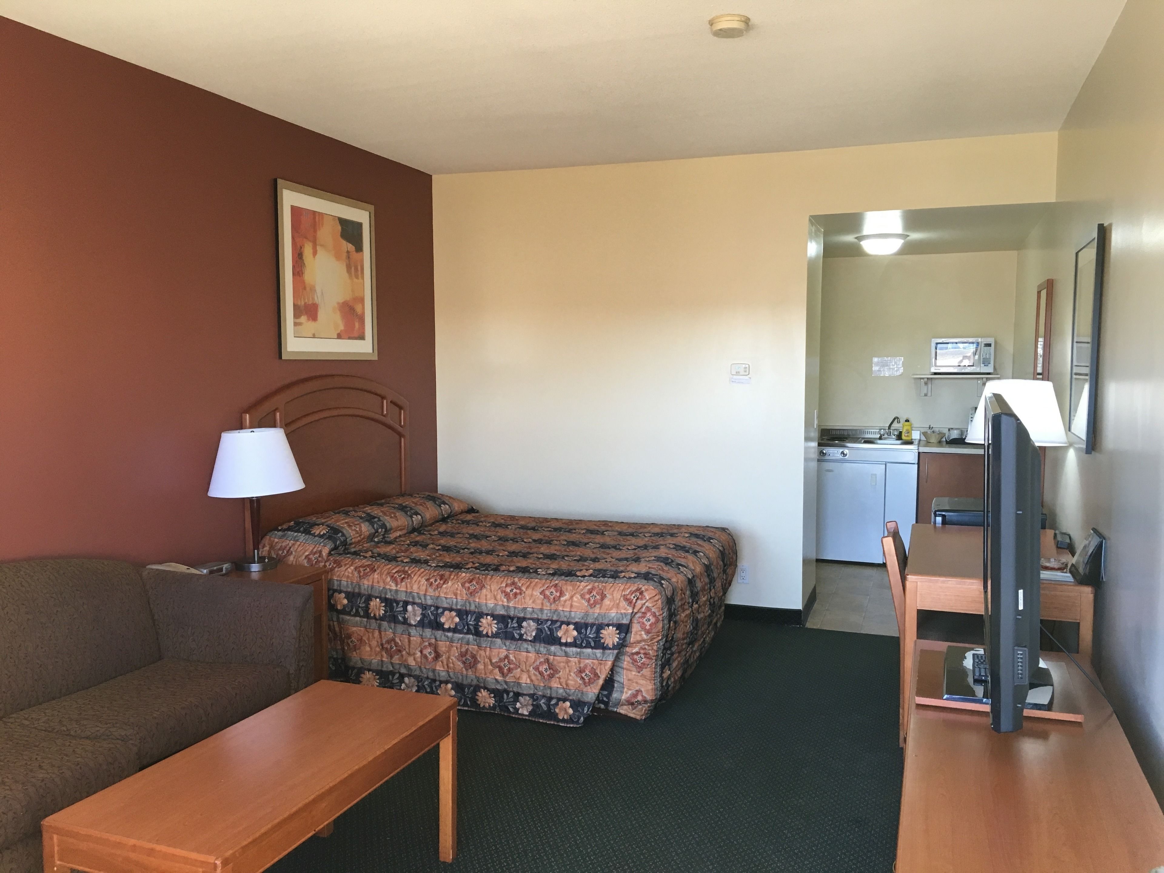 A compact room with a bed, a small desk, a chair, a sofa, and a kitchenette area.
