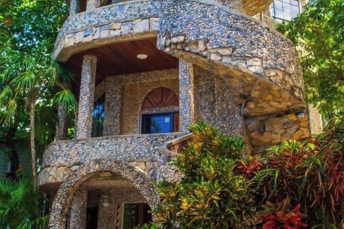 A multi-level stone building with balconies and arched windows, surrounded by lush greenery.