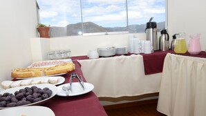 Un bufé de desayuno con diversas opciones de comida y bebidas, una ventana con vistas a la montaña y una mesa con mantel rojo.