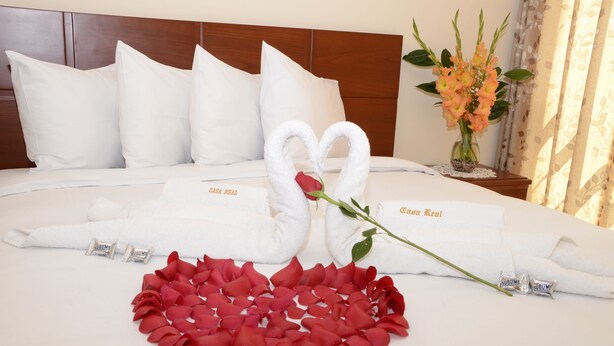 Una cama bien tendida con almohadillas blancas, un arreglo con pétalos de rosa roja en forma de corazón, y dos toallas blancas moldeadas como cisnes con una rosa roja en medio.