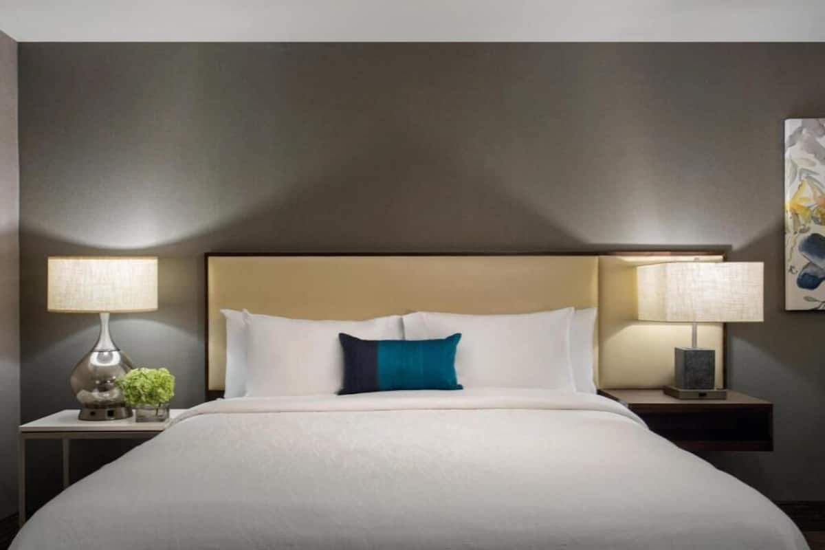 Quarto de hotel com uma cama grande, duas mesas de cabeceira com luminárias, uma mesinha de apoio e uma obra de arte pendurada na parede.