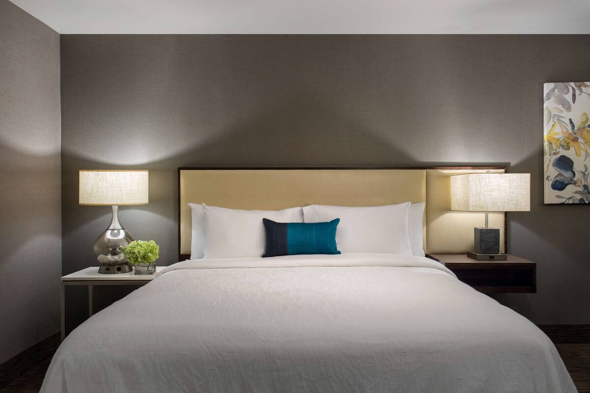 Cama bem arrumada com lençóis brancos, duas mesas de cabeceira com luminárias e uma obra de arte pendurada na parede.