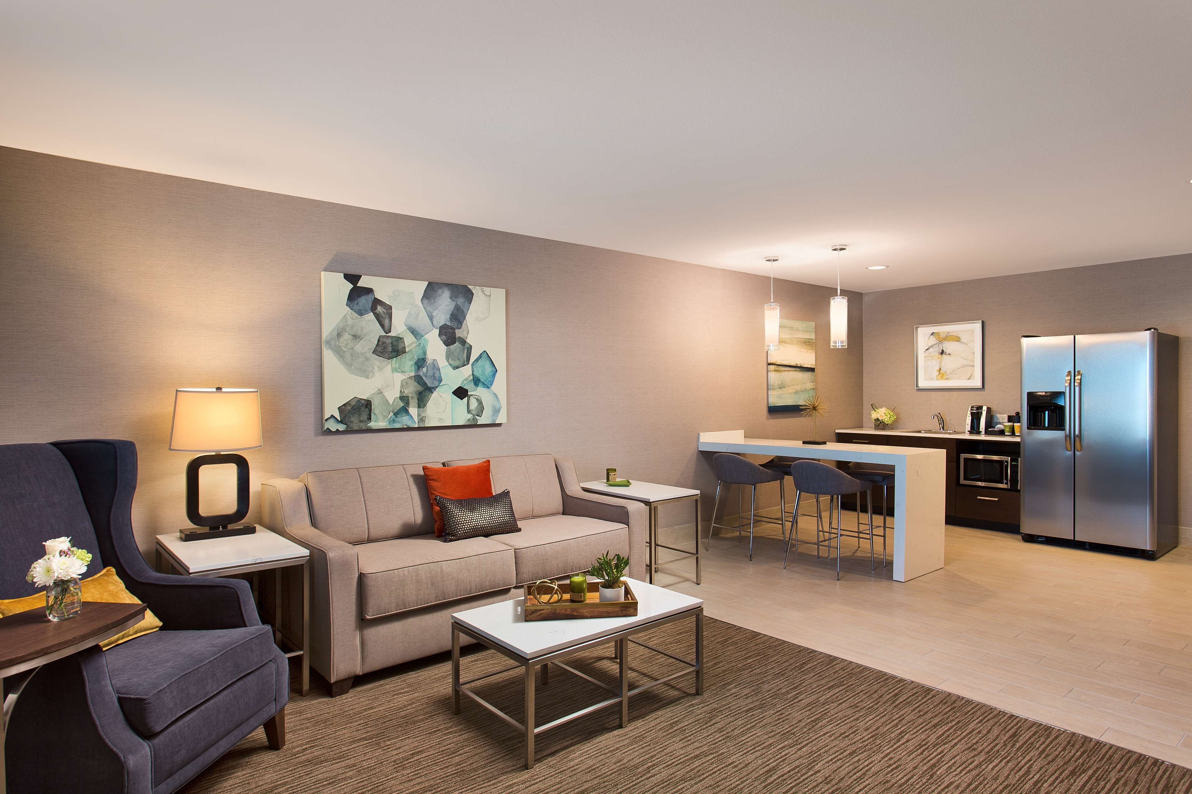 A modern living room with a sofa, coffee table, and a small kitchen area with a refrigerator and bar stools.