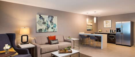 A modern living room with a sofa, coffee table, and a small kitchen area with a refrigerator and bar stools.