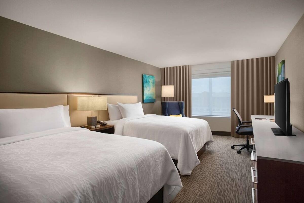 Quarto de hotel com duas camas, uma escrivaninha, uma cadeira, uma televisão e uma janela com cortinas.