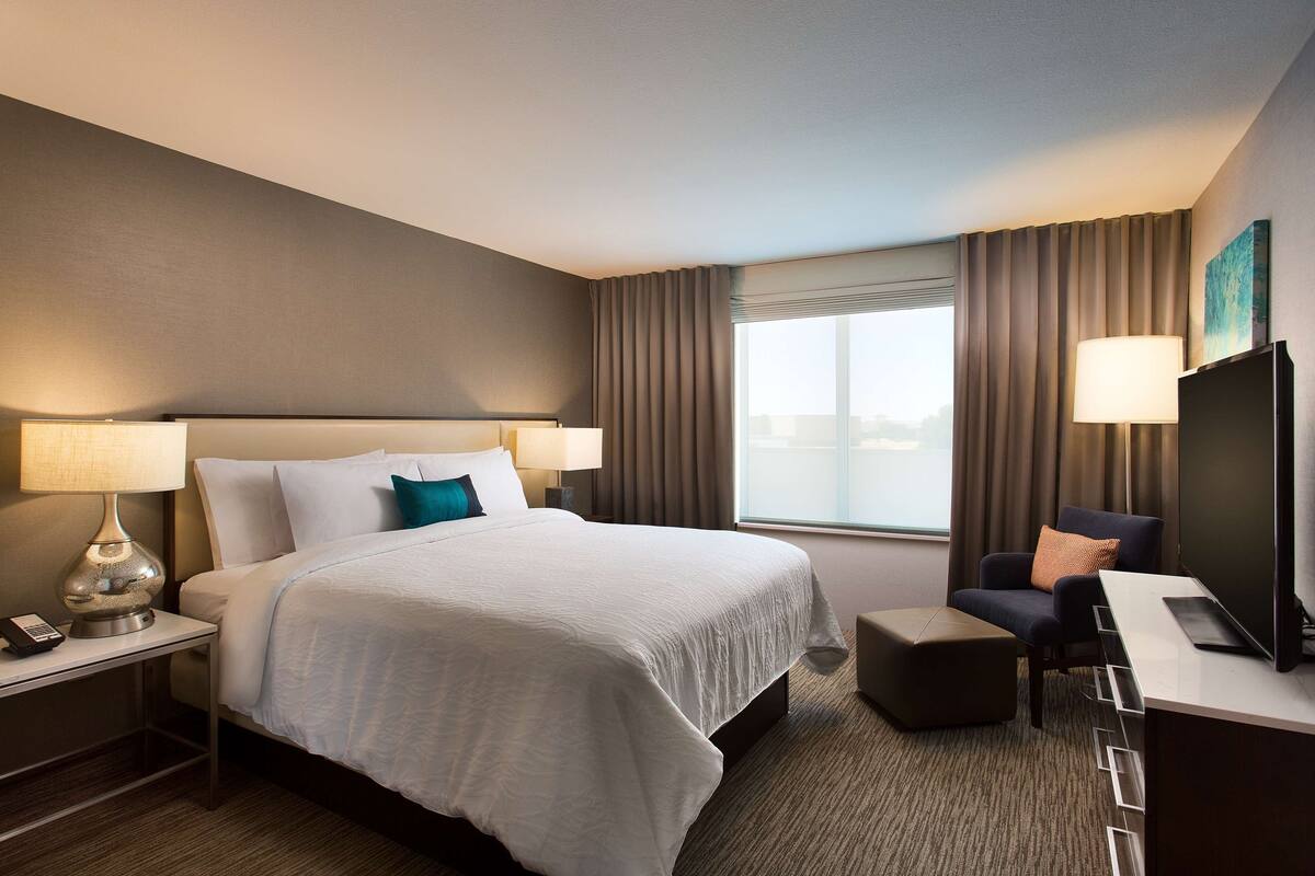 Quarto de hotel com uma cama grande, uma escrivaninha com cadeira, uma luminária e uma televisão.