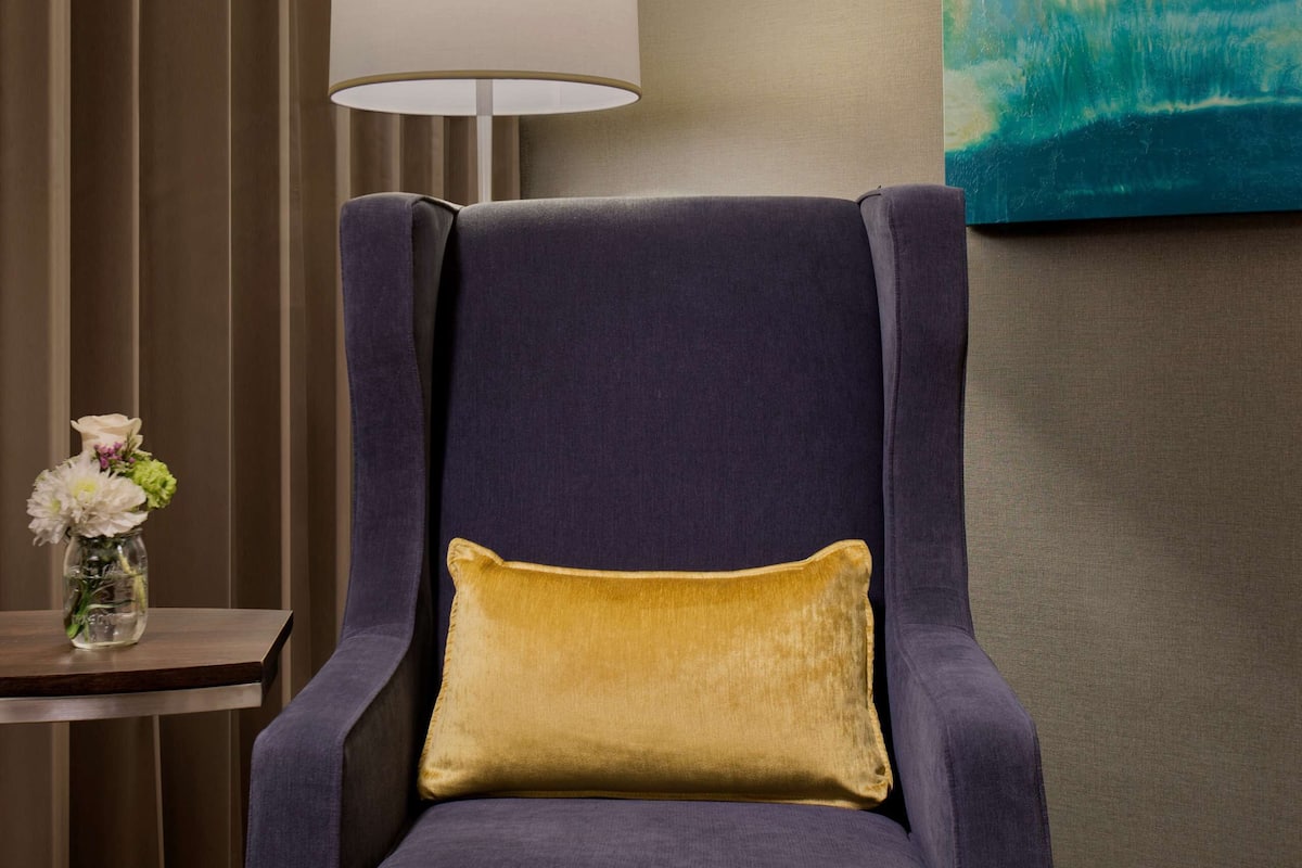 Quarto com poltrona azul escura, almofada dourada, luminária branca, um quadro e uma mesinha com um vaso de flores.