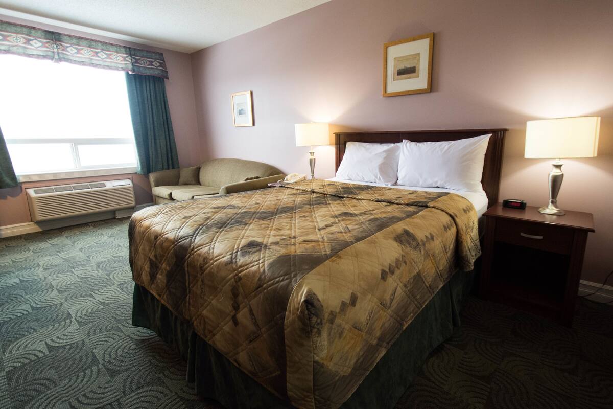 A hotel room with a large bed, two bedside lamps, a chair, a window with curtains, and a framed picture on the wall.
