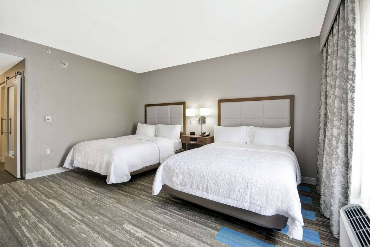 A hotel room with two beds, a nightstand with a lamp, a window with curtains, and a door leading to another room.
