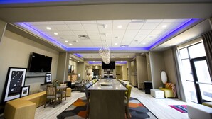 A modern hotel lobby with a long dining table, comfortable seating, a flat-screen TV, and a large window offering a view. - Hampton Inn Blue Ash/Cincinnati (Cincinnati)