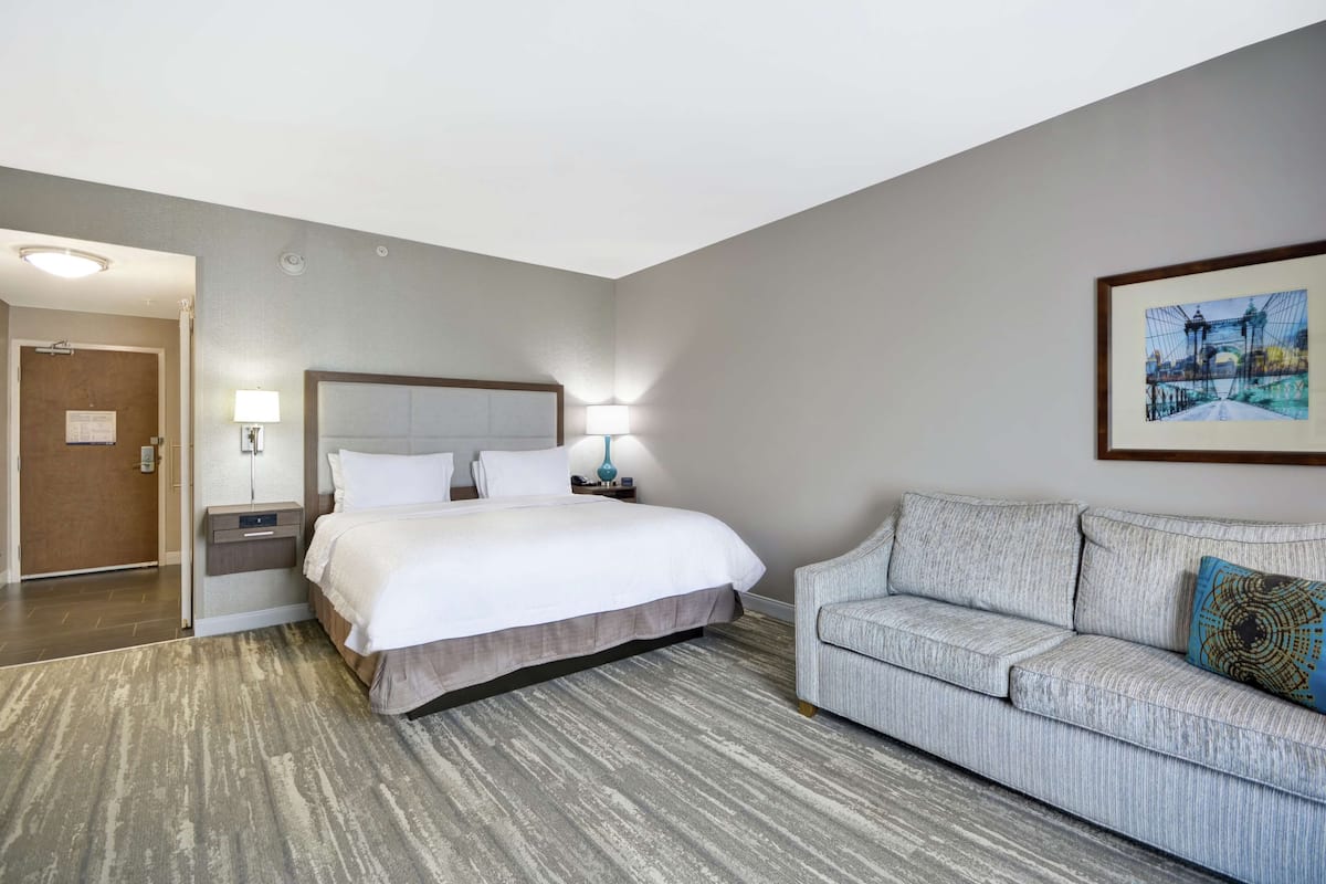 A hotel room with a bed, a nightstand, a lamp, a sofa, and a framed picture on the wall.