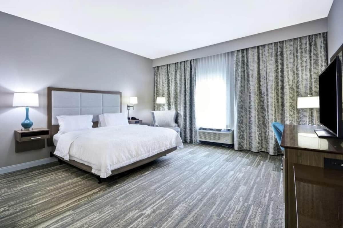 A hotel room with a bed, bedside tables, a TV, and a large window with curtains.