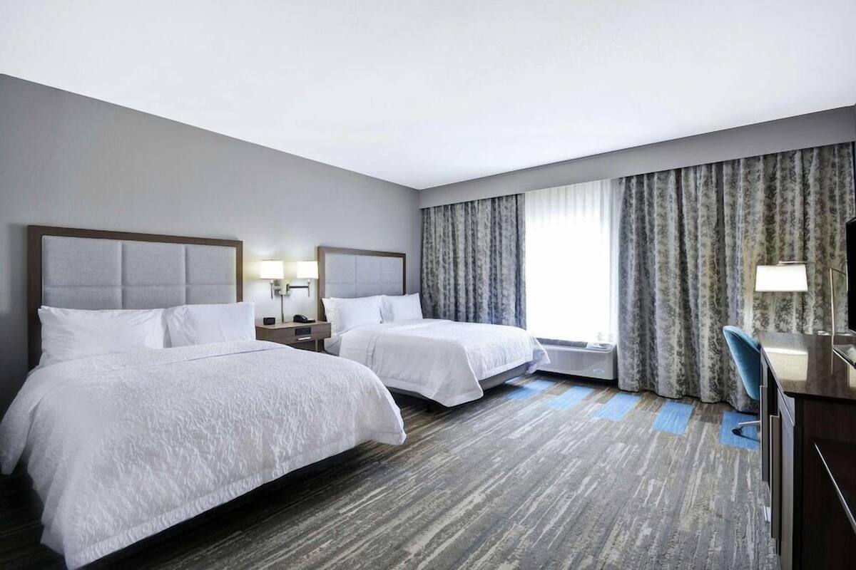 A hotel room with two beds, a desk, a chair, and a large window with curtains.