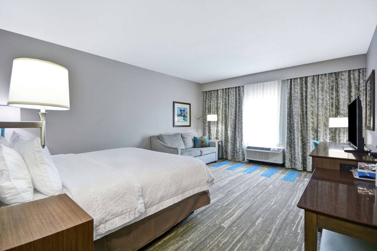 A hotel room with a large bed, a desk, a chair, a television, and a window with curtains.