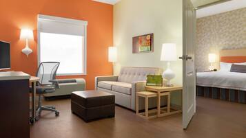 A hotel room with a desk, office chair, sofa, ottoman, bedside tables with lamps, a bed, and a window.