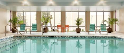 Una piscina interior con agua azul cristalina, rodeada de sillones de descanso y plantas en macetas.