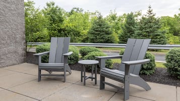 Two Adirondack chairs and a small round table on a concrete patio with a view of trees and a guardrail.