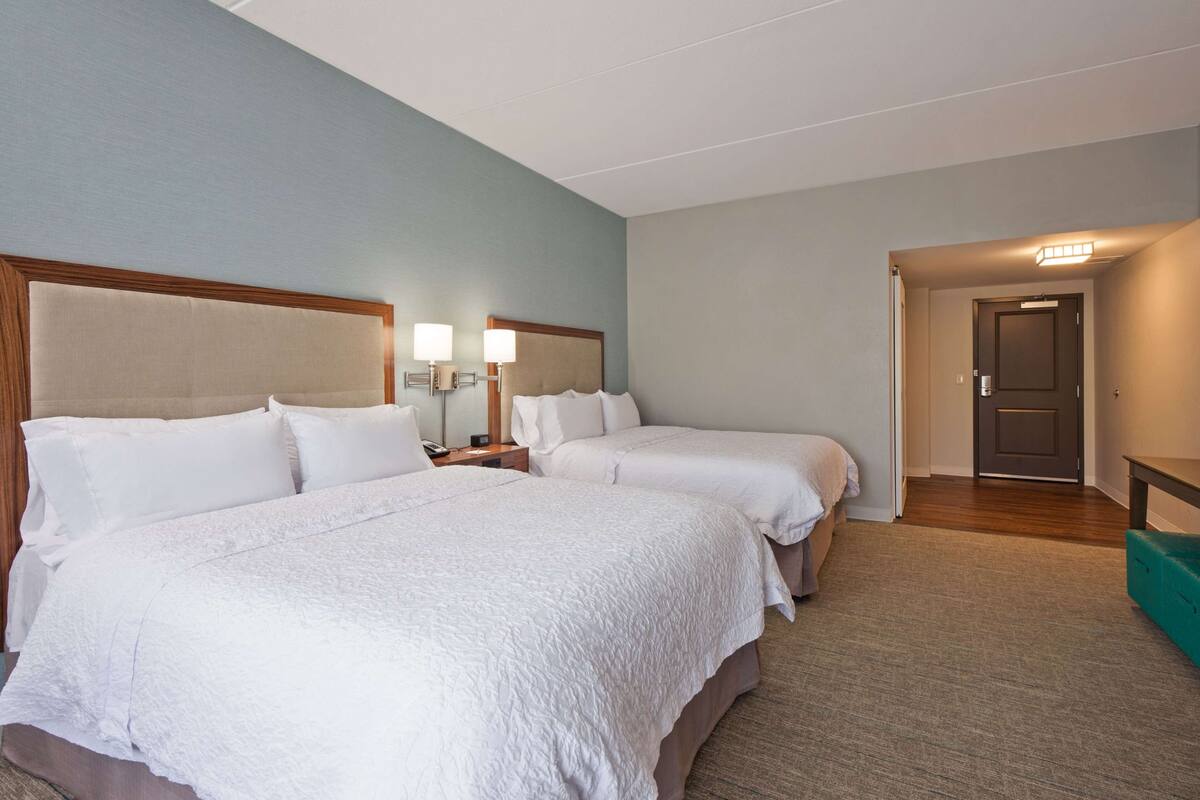 Ein Hotelzimmer mit zwei Betten, einem hölzernen Kopfteil, einem Nachttisch mit Lampe und einem Teppichboden.
