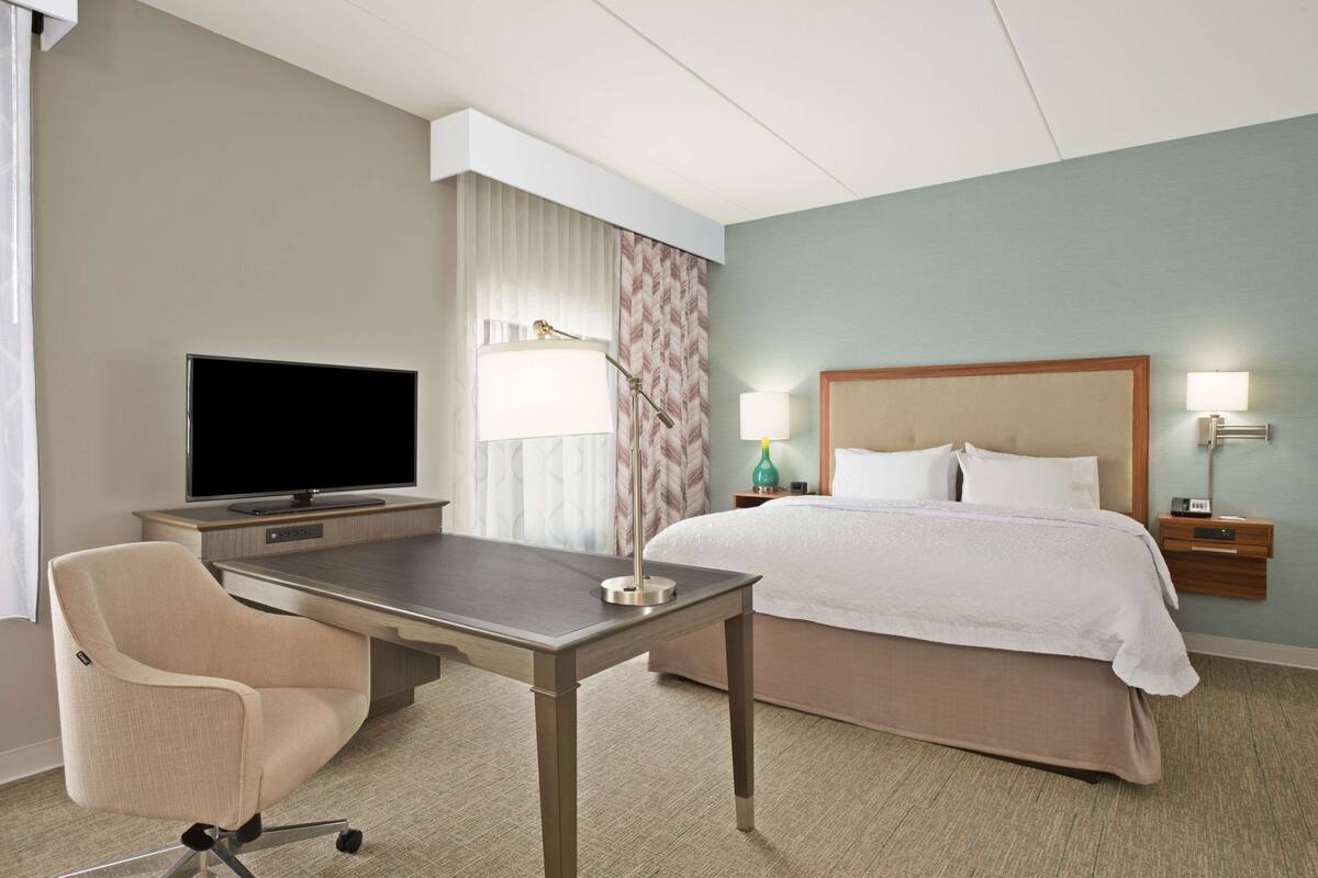 Ein Hotelzimmer mit einem Bett, einem Schreibtisch mit Fernseher, einem Sessel und einem Nachttisch mit Lampen.