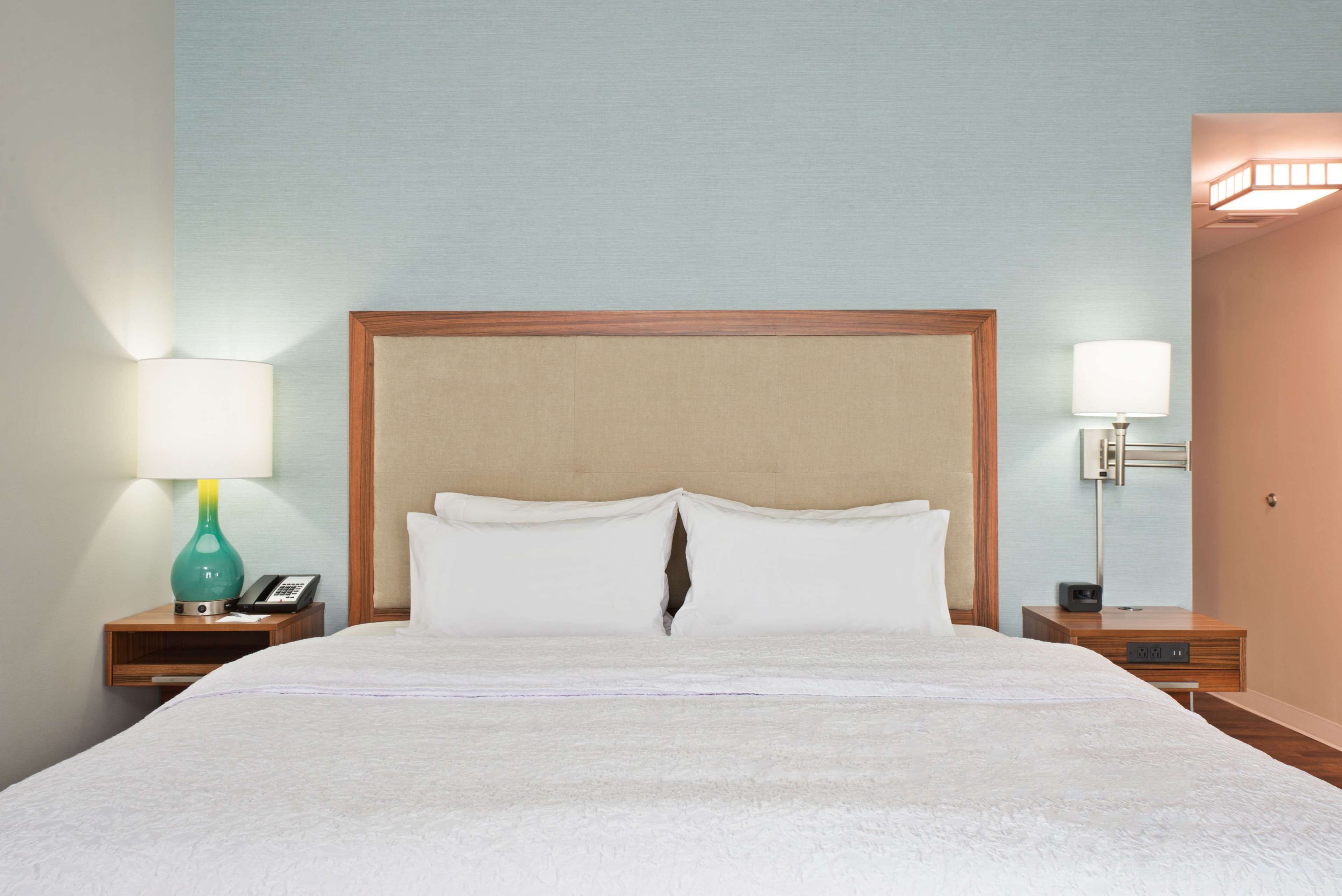 Cama bem arrumada com cabeceira de madeira, duas mesas de cabeceira com abajures e luminária fixada na parede.