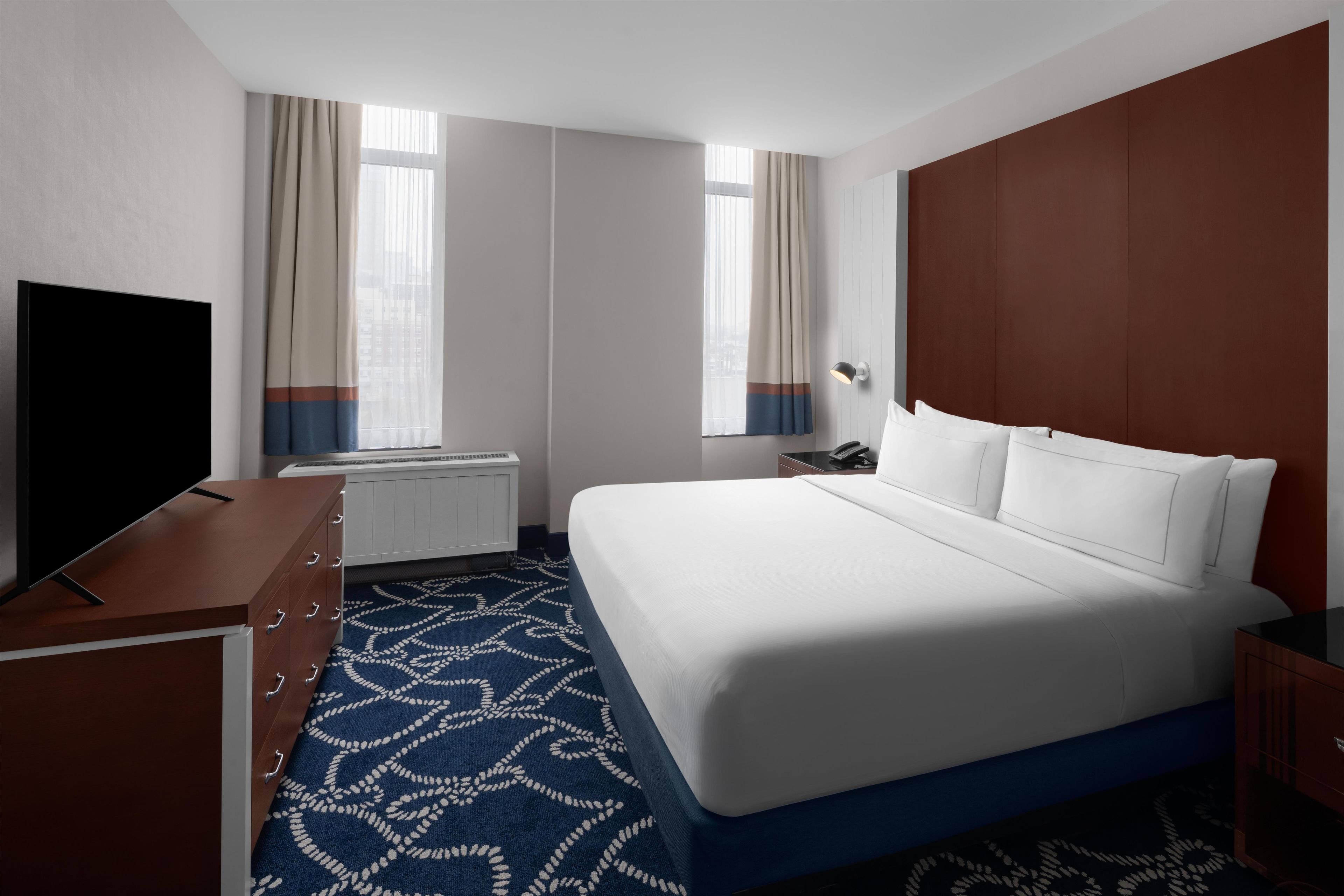 A hotel room with a large bed, a television on a wooden dresser, a desk, and a patterned carpet.