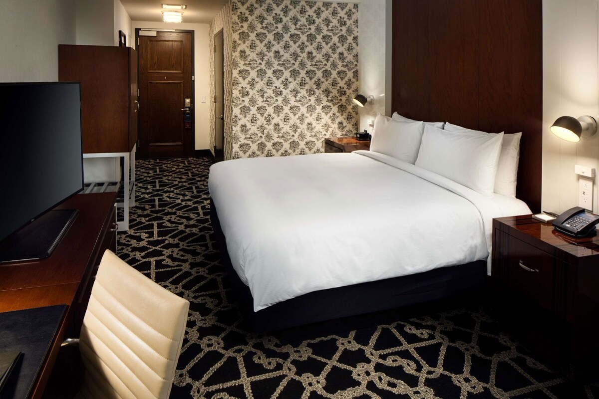 A hotel room with a bed, a desk with a TV, a telephone, and a patterned carpet.