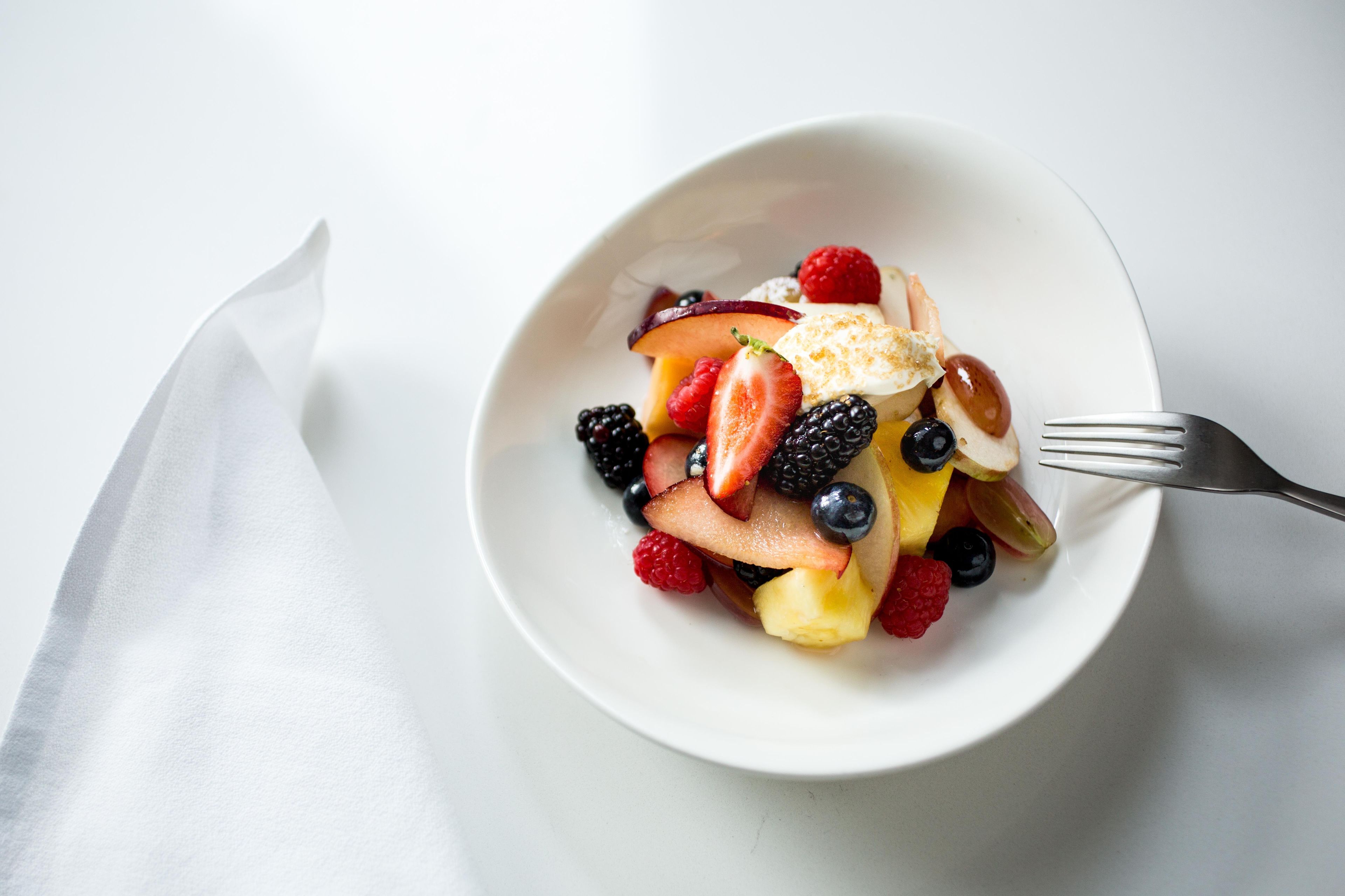 Un tazón blanco lleno de frutas variadas y un pedazo de pastel, acompañado de un tenedor sobre una superficie blanca.